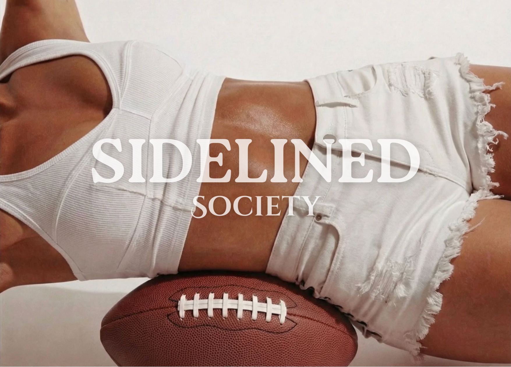 A person lying on a white surface wearing a white ribbed sports bra and distressed white denim shorts with frayed hems, with a brown football resting beneath the shorts. Large white text across the center reads 'SIDELINED SOCIETY'.